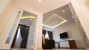 Una sala moderna con un televisor de pantalla plana montado en la pared, un sofá de color oscuro, un mueble de madera y una puerta de vidrio que da a otra habitación.