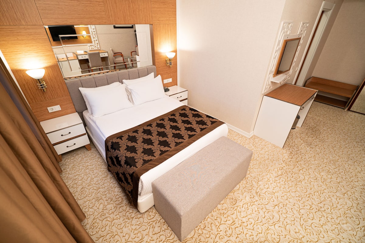 A hotel room with a bed, bedside tables, a bench, a mirror, and a TV.