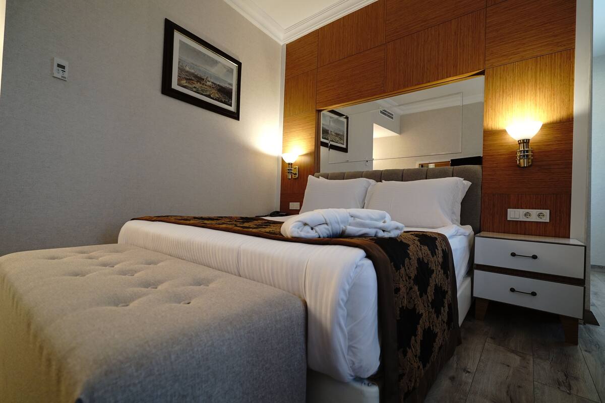 A hotel room with a large bed, a bedside table, a nightstand, a mirror, and two wall-mounted lights.