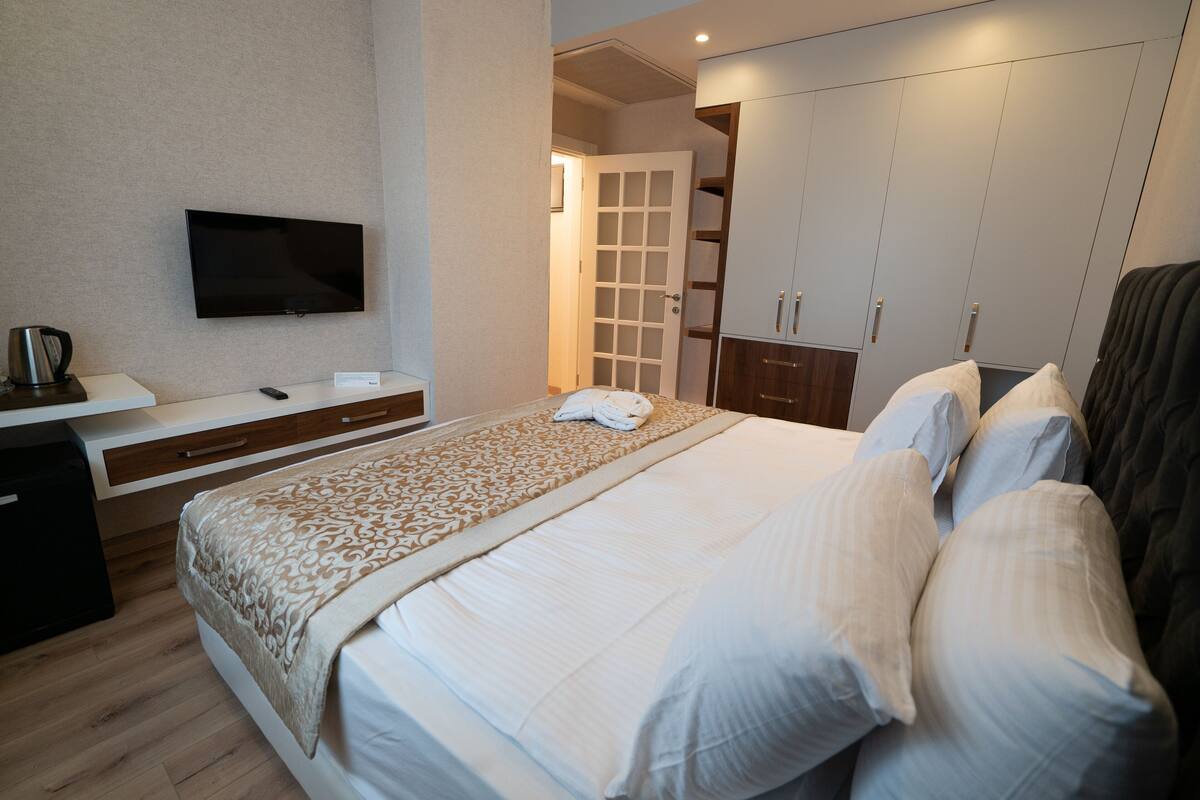 A hotel room with a large bed, a flat-screen TV mounted on the wall, a bedside table with a kettle, and a wardrobe with multiple doors.