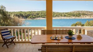 Et træbord med stole og en bænk på en balkon med udsigt over et kystlandskab med træer og en strand.