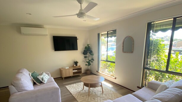 A spacious living room with a large sofa, a smaller sofa, a coffee table, and a ceiling fan. There is a flat-screen TV mounted on the wall, a potted plant, and a window with a view of greenery outside.