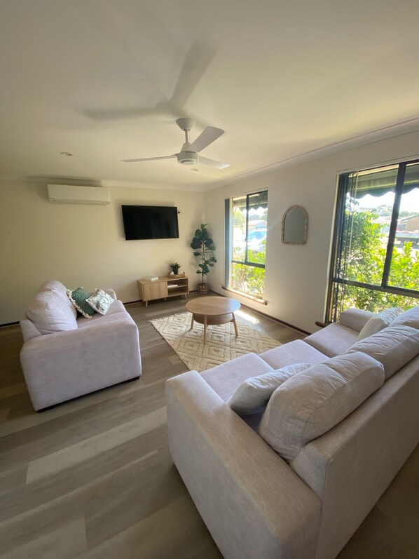 A spacious living room with a large sofa, a smaller sofa, a coffee table, and a ceiling fan. There is a flat-screen TV mounted on the wall, a potted plant, and a window with a view of greenery outside.