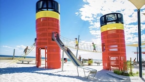 Two cylindrical structures with ladders, a slide, and a rope bridge on a sandy beach.