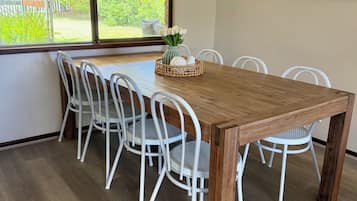 A dining area with a wooden table and white chairs, a window with a view of greenery, and a painting on the wall.