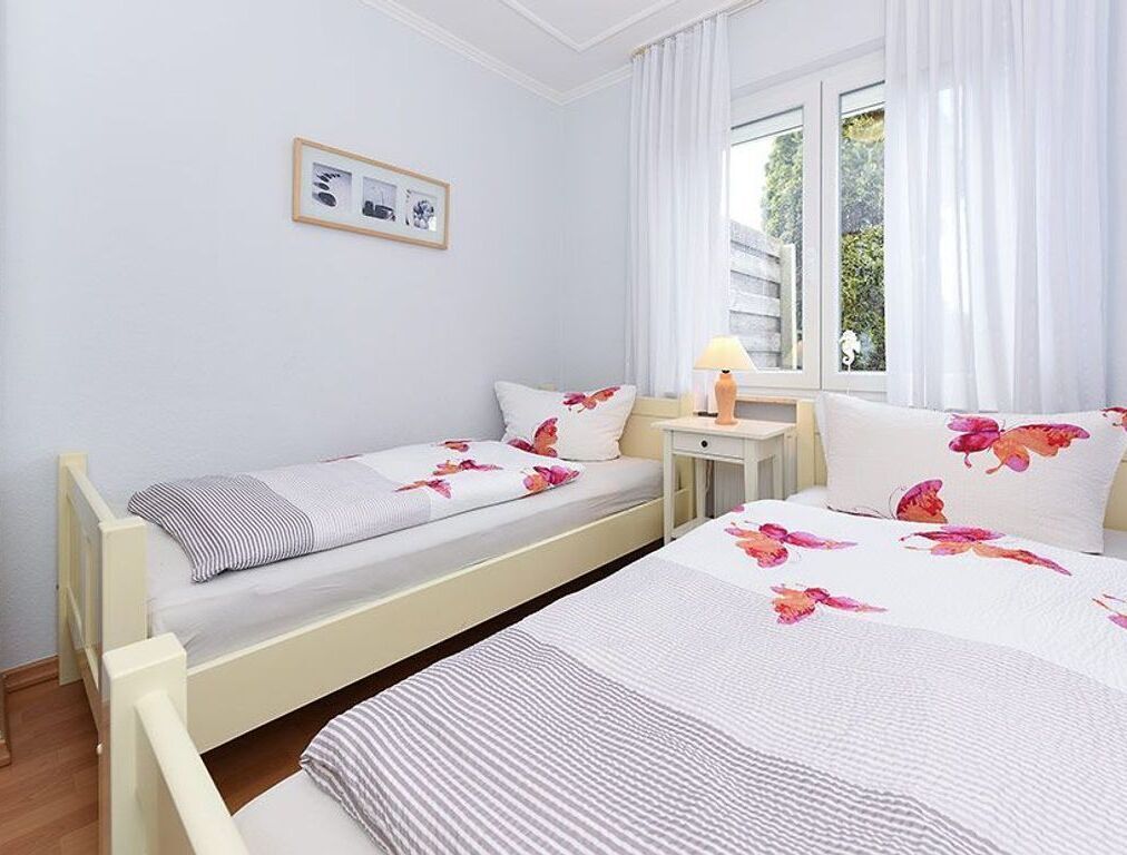 Una cama doble con ropa de cama floral, una mesita de noche con lámpara, un cuadro pequeño en la pared y una ventana con cortinas.