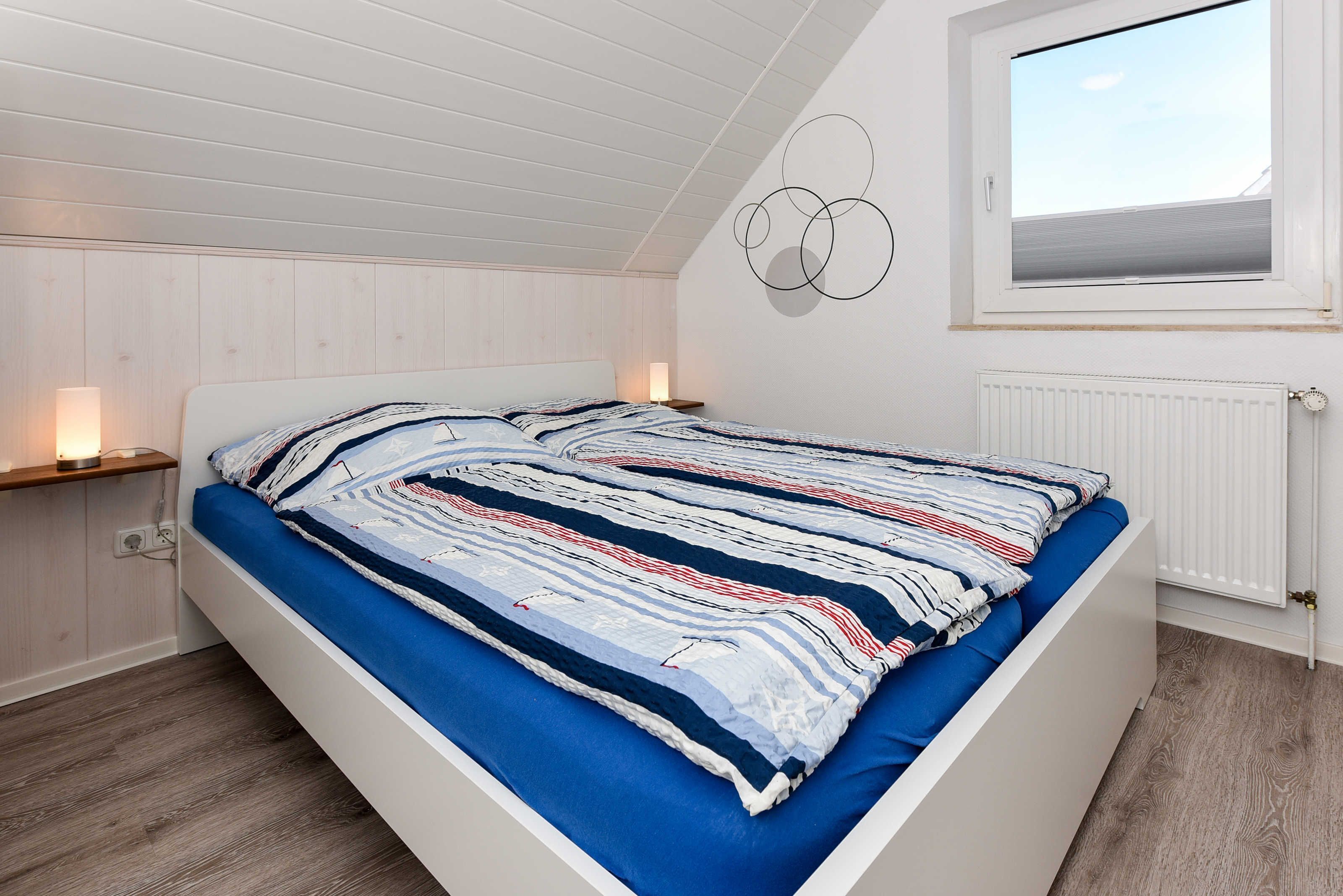 Un dormitorio con techo inclinado, una cama con colcha a rayas, una lámpara fijada a la pared y una ventana con vistas al cielo.