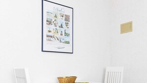 Un petit coin repas avec une table blanche et deux chaises blanches dotées de coussins noirs. Un tableau encadré est accroché au mur, au-dessus de la table.