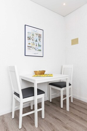 Een kleine eethoek met een witte tafel en twee witte stoelen met zwarte kussens. Daarboven hangt een lijst met een afbeelding aan de muur.