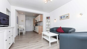 Un salon moderne avec un canapé gris, une table basse blanche et un téléviseur posé sur un meuble. On aperçoit, au fond, une cuisine avec des meubles en bois et une étagère blanche remplie de livres.