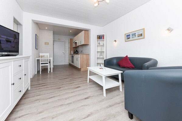 Ein modernes Wohnzimmer mit einem grauen Sofa, einem weißen Couchtisch und einem Fernseher auf einem Schrank. Im Hintergrund ist eine Küche mit Holzschränken und einem weißen Bücherregal zu sehen.