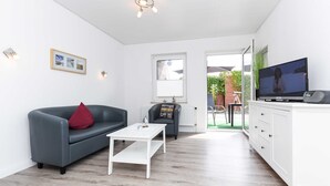 Un salon moderne avec un canapé gris, une table basse blanche, un téléviseur et une vue sur un jardin, visible à travers une porte vitrée.