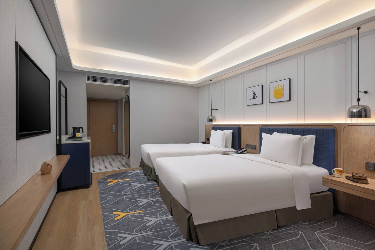 A hotel room with two beds, a desk, a TV, and a small kitchenette.