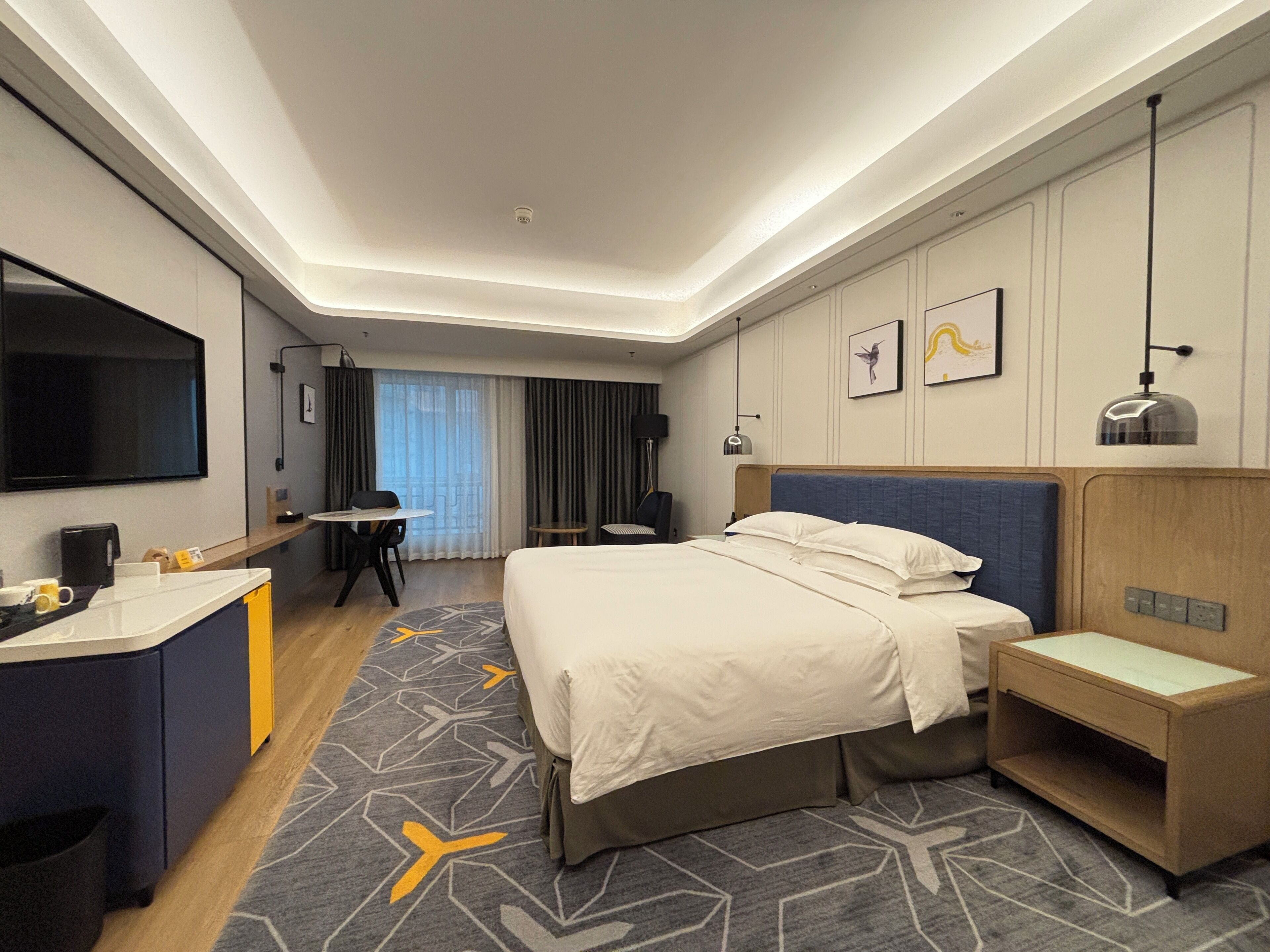 A hotel room with a bed, bedside table, desk, and chair. There is a TV mounted on the wall, a window with curtains, and a patterned carpet.