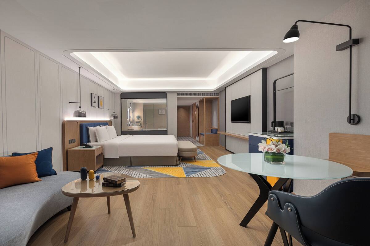 A modern hotel room with a bed, a sofa, a small table, a TV, and a bathroom visible through an open door.