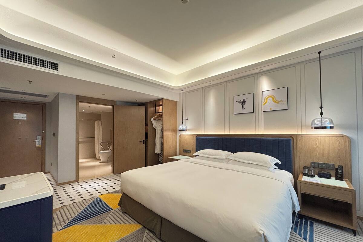 A modern hotel room with a large bed, bedside tables, a desk, and a bathroom visible through an open door.