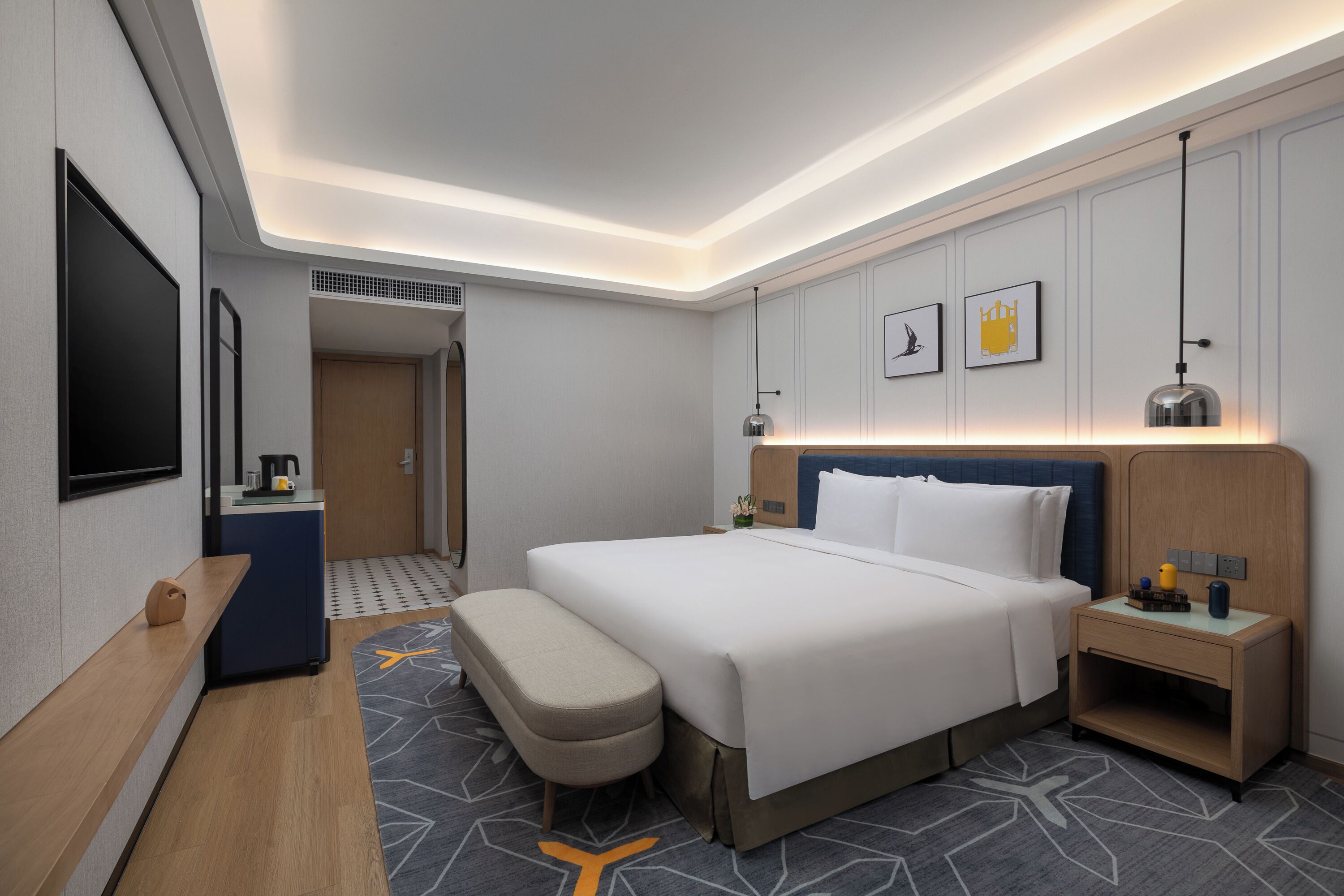 A modern hotel room with a bed, bedside table, bench, and a small desk with a kettle.