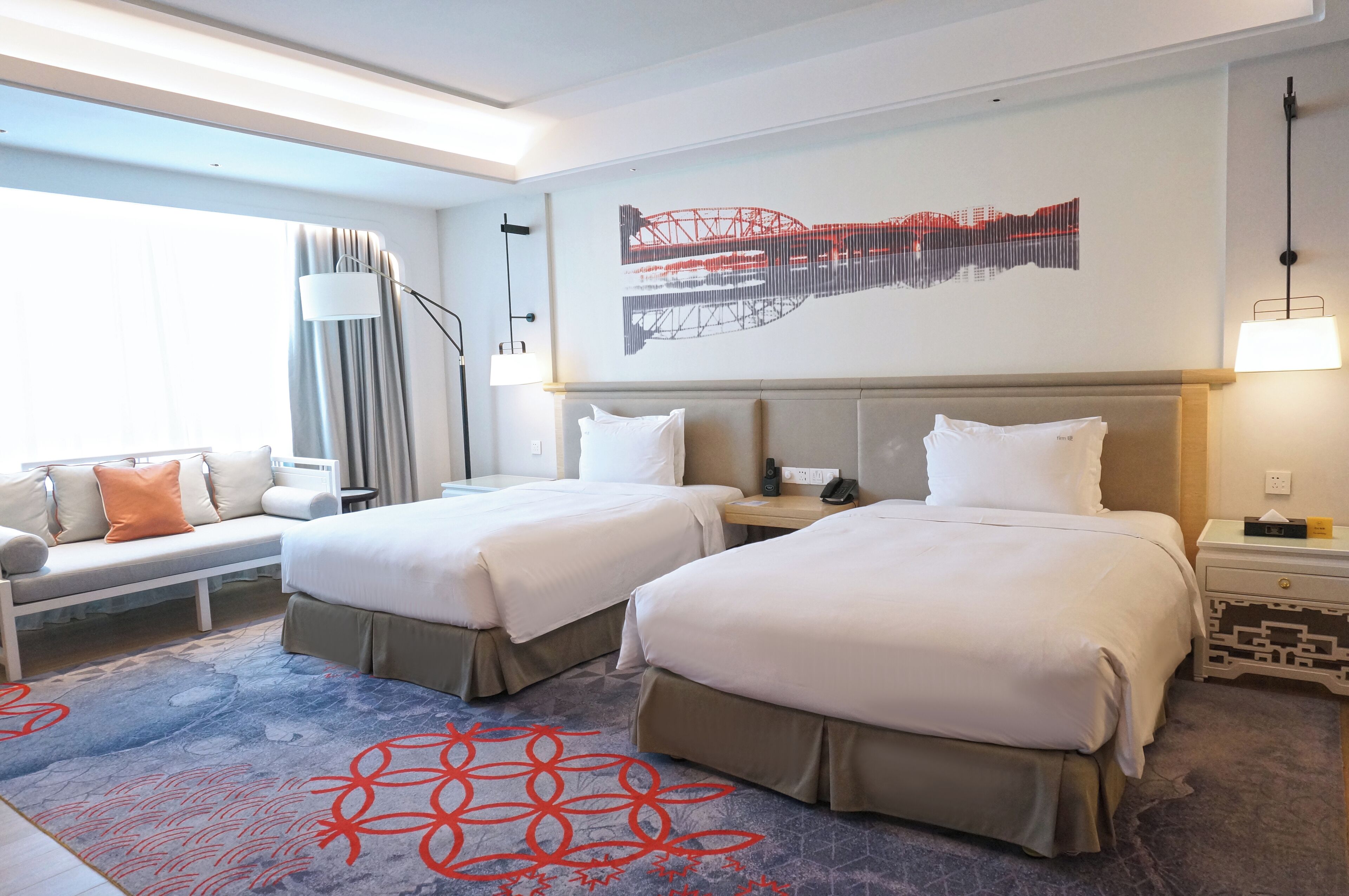 A hotel room with two beds, a bench, a desk, and a wall-mounted artwork.