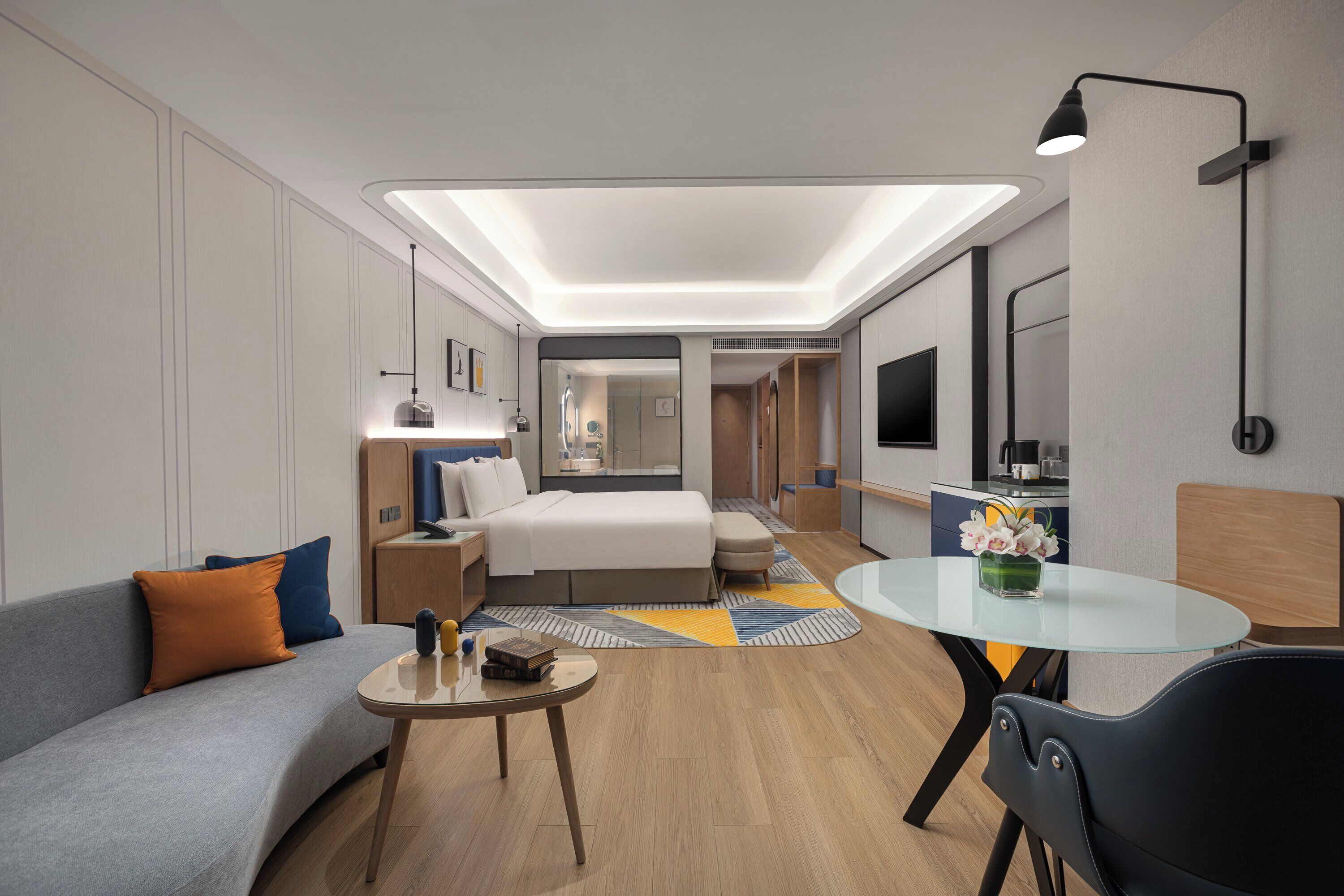 Ein modernes Hotelzimmer mit Bett, Sofa, einem kleinen Tisch, Fernseher und einem durch eine Glastrennwand einsehbaren Badezimmer.