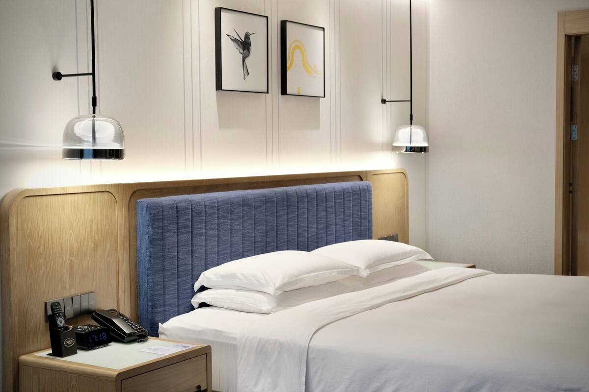 A hotel room with a wooden headboard, a bed with white linens, a bedside table with a phone and a lamp, and two framed artworks on the wall.