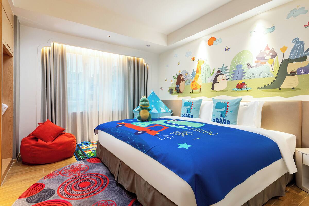 A child-friendly hotel room with a bed, a red bean bag chair, a play mat, and a wall mural.