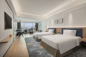 A hotel room with two beds, a desk, and a large window offering a city view.