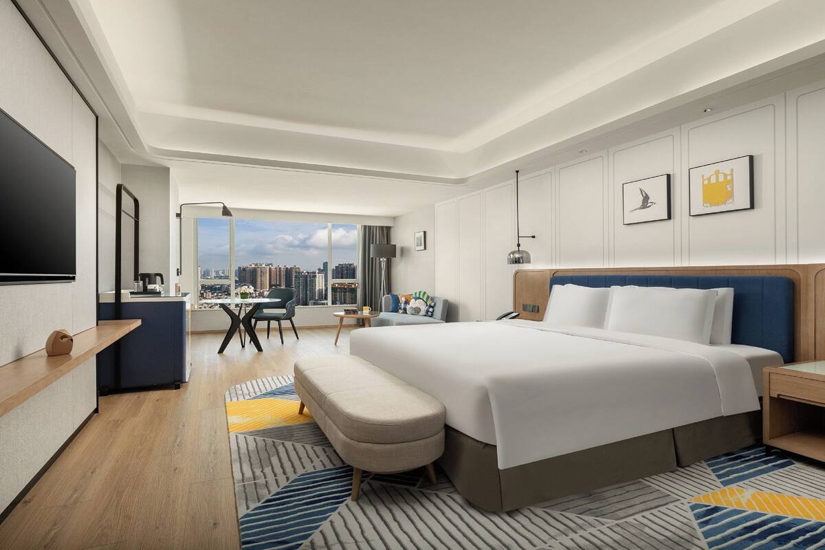 A modern hotel room with a large bed, a desk, a chair, and a view of the city.