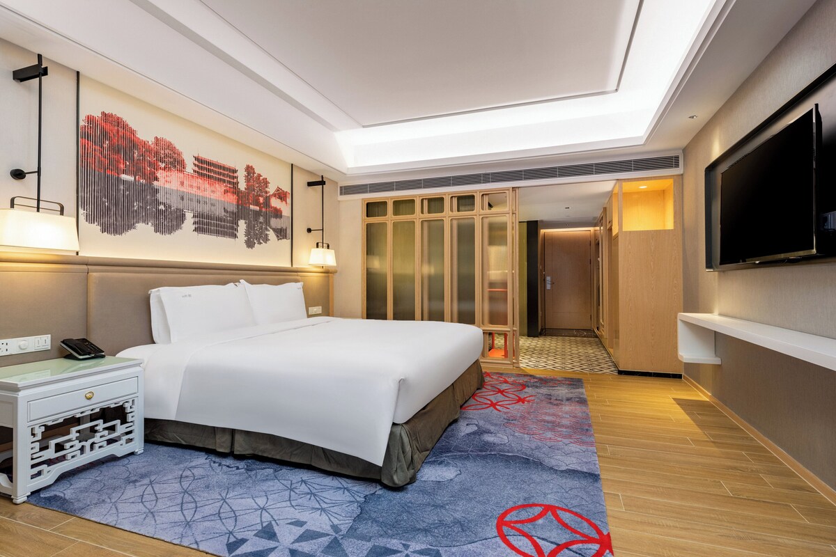 Una habitación de hotel moderna con una cama grande, un televisor de pantalla plana, un escritorio con lámpara y un adorno de pared decorativo.