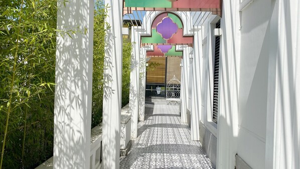 Un pasillo estrecho, con paredes blancas y baldosas de piso estampadas, que conduce a la entrada de un edificio con un arco decorativo.