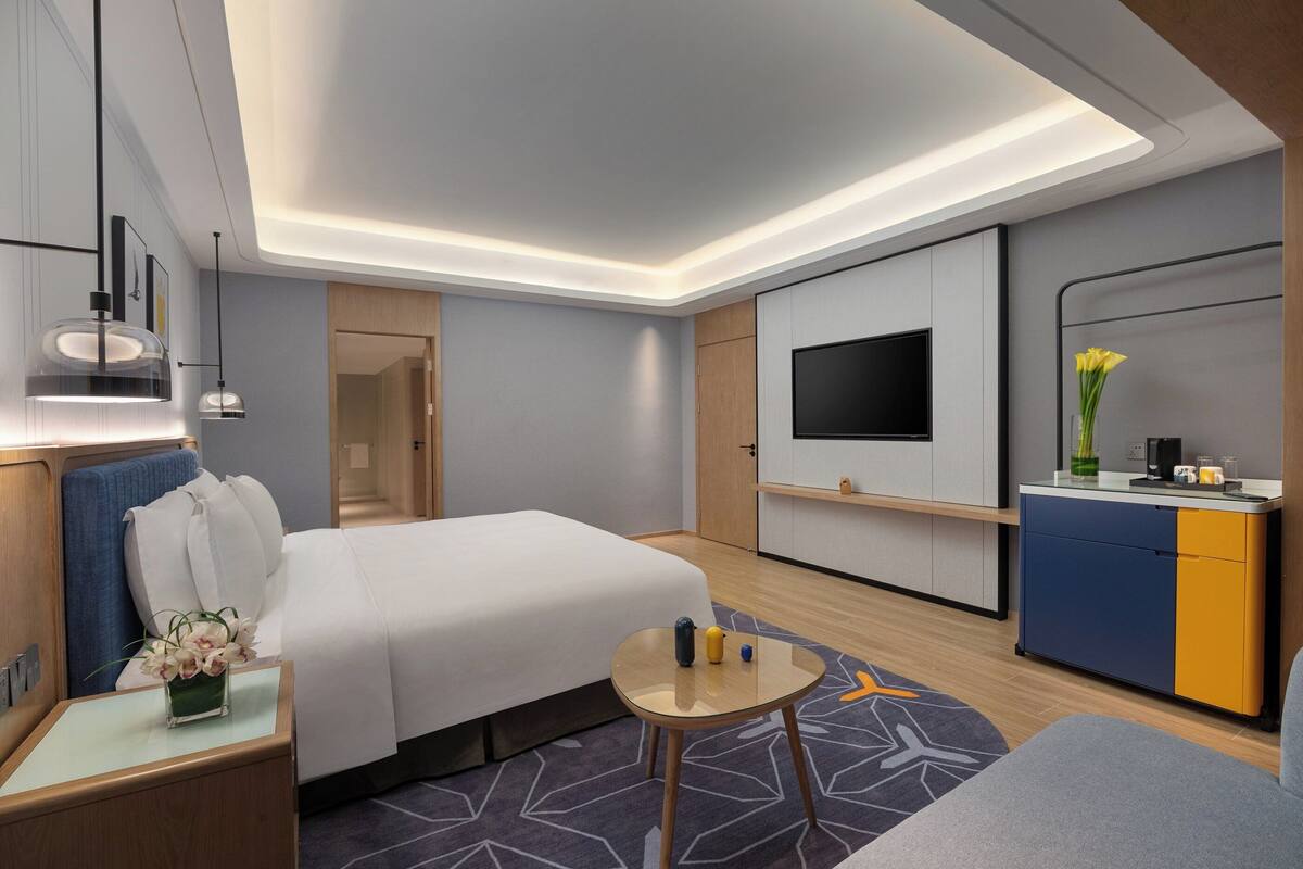A modern hotel room with a bed, bedside table, lamp, TV, and a small desk area.