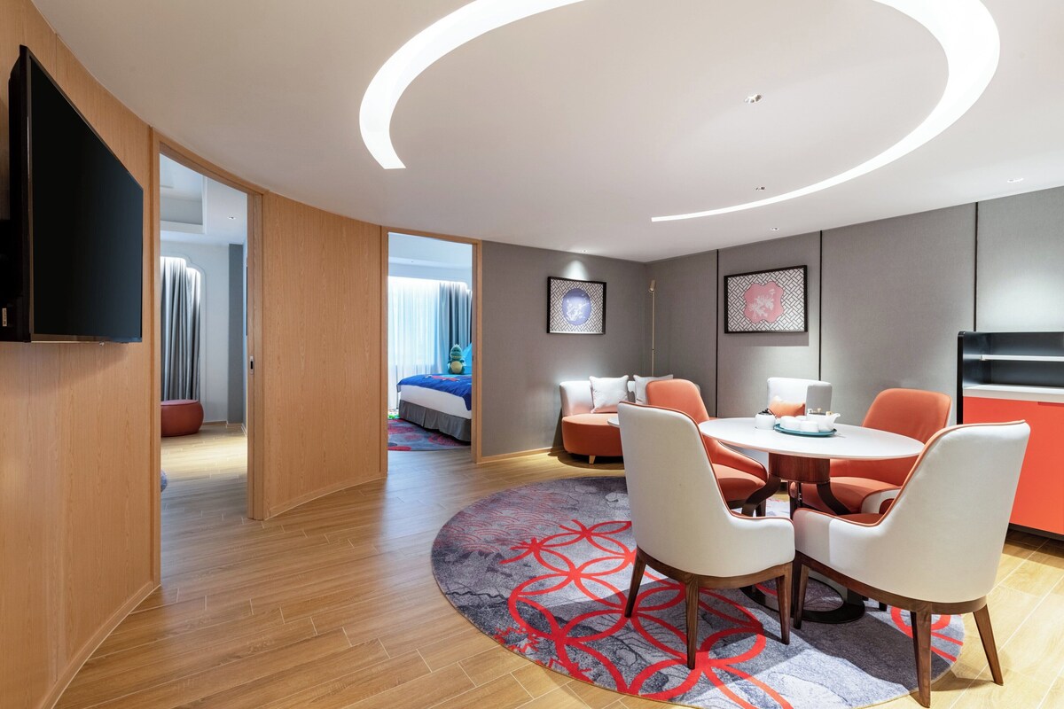 Una habitación de hotel moderna con una mesa de comedor redonda, sillas blancas y naranjas, un televisor de pantalla plana en la pared y un gran ventanal con cortinas.