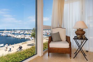 A room with a view of a marina, a wicker chair with a cushion, a table with a lamp, and sheer curtains.