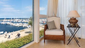 Ein Zimmer mit Blick auf eine Marina, ein Korbstuhl mit Kissen, ein Tisch mit einer Lampe und transparente Vorhänge.