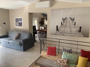 Un salon avec un canapé gris, un coin repas avec des chaises en bois, une cuisine à l’arrière-plan et une grande œuvre d’art représentant un pont en bois.