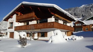 A two-story chalet with a wooden balcony, snow-covered roof, and a mountain backdrop. - Ritter Anna - Haus Panorama - Ferienwohnung (Schoppernau)