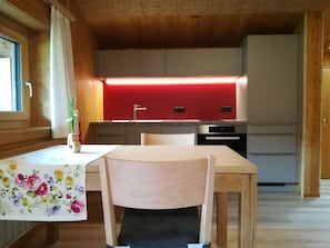 A wooden interior with a dining table, chairs, a red-lit kitchen, and a window with curtains. - Ritter Anna - Haus Panorama - Ferienwohnung (Schoppernau)