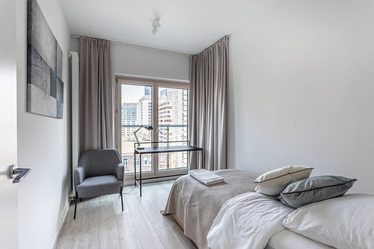 A modern hotel room with a bed, a desk, a chair, and a large window with a city view.