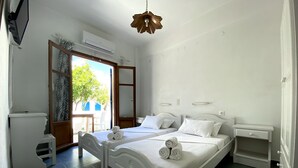 A bedroom with two beds, a small table, a chair, and a window with a view of greenery. - Helliniko Studios - Adults Only  (Paros)