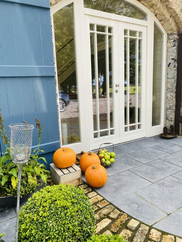 A house with a blue exterior, a white door with glass panels, and a stone wall. There are pumpkins and a basket of apples on the porch.
