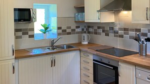 A modern kitchen with white cabinetry, a built-in microwave, a kettle, and a stove. - Le Petit Maison - La Relierie (Gavray)