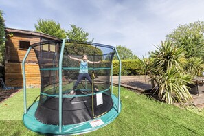 A trampoline with a safety net enclosure in a backyard with a wooden house and green lawn.