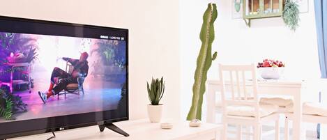 A living room with a flat-screen TV on a white stand, a cactus plant, a wooden chair, a dining table with chairs, and decorative fish on the wall.