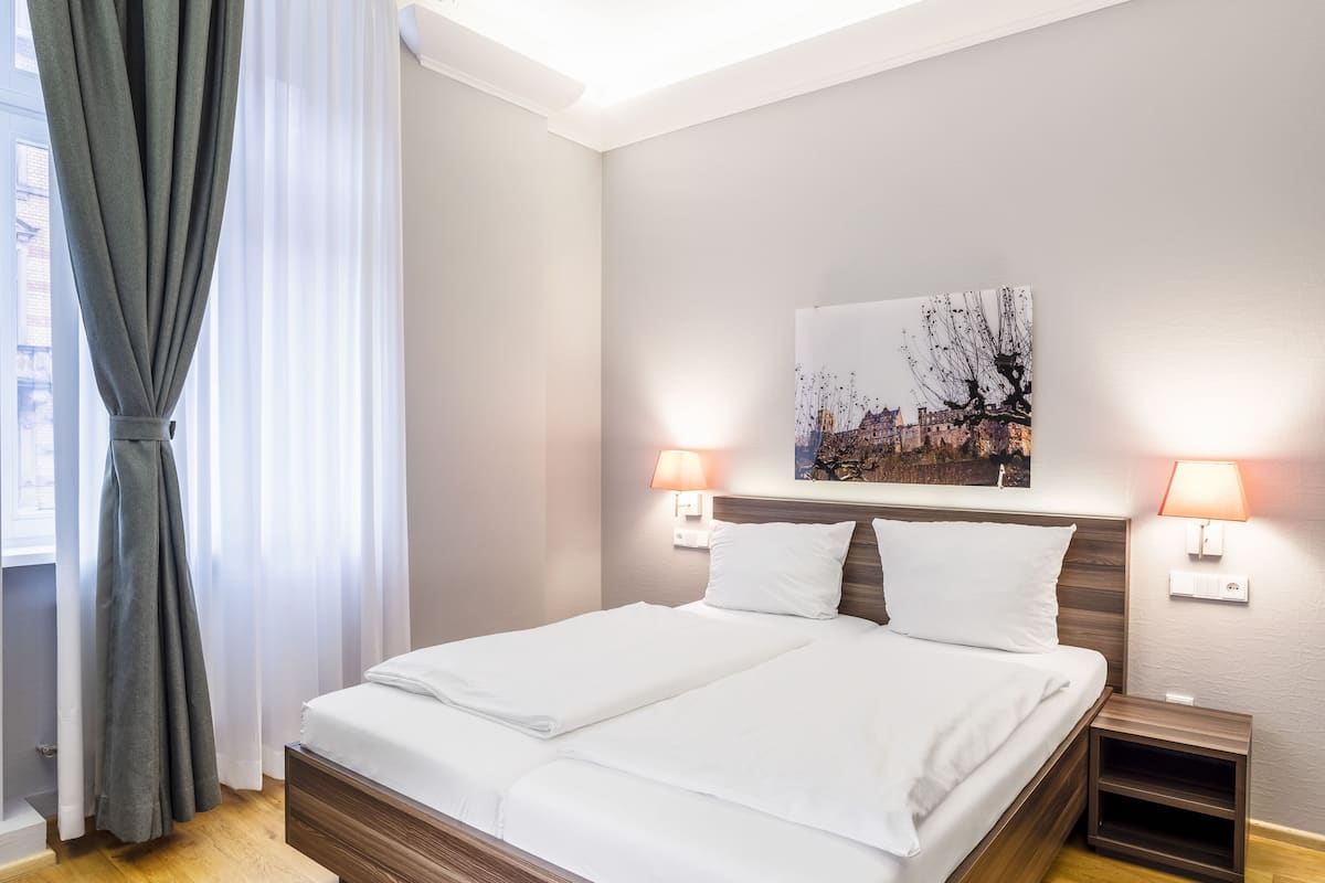 Une chambre à coucher moderne avec un grand lit, une tête de lit en bois, deux lampes de chevet et un tableau représentant un paysage urbain accroché au mur.