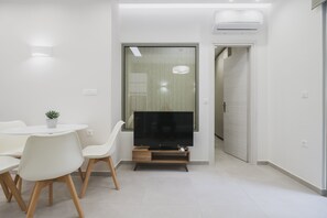 A modern living room with a flat-screen TV on a wooden stand, white chairs, a small potted plant, and a sliding glass door leading to another room.