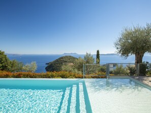 A clear blue sky, a swimming pool with clear water, and a view of the sea and distant mountains.