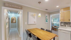 Une cuisine avec une table en bois, des chaises et un luminaire suspendu. Au-dessus de la porte, on peut lire une inscription : « MAISON DE PLAGE ».