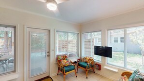 Une pièce avec un téléviseur fixé au mur, deux fauteuils en osier avec des coussins, une petite bibliothèque et une porte avec des stores.