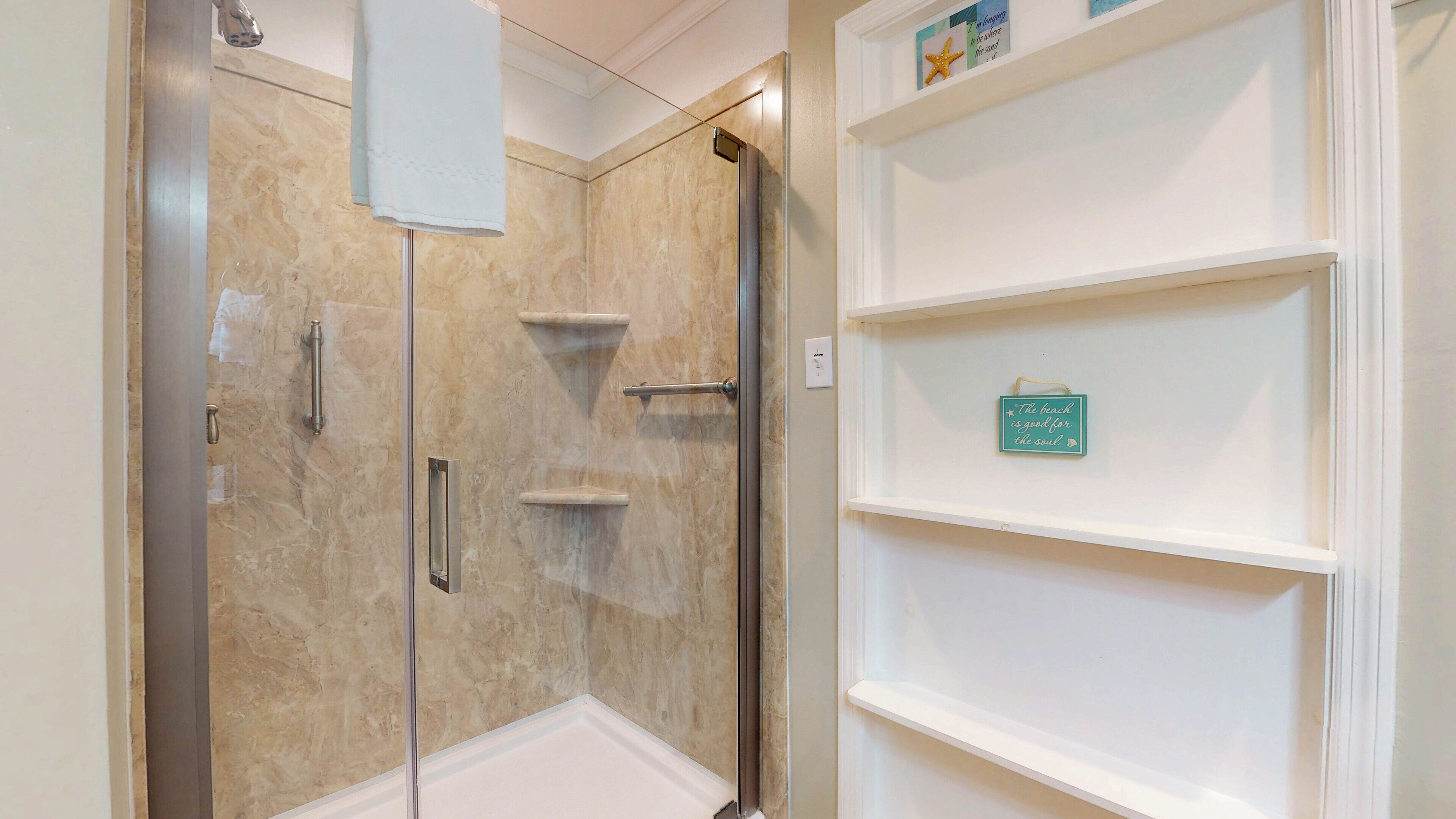 A bathroom with a glass shower enclosure, a white sink, and a shelf with a decorative sign.