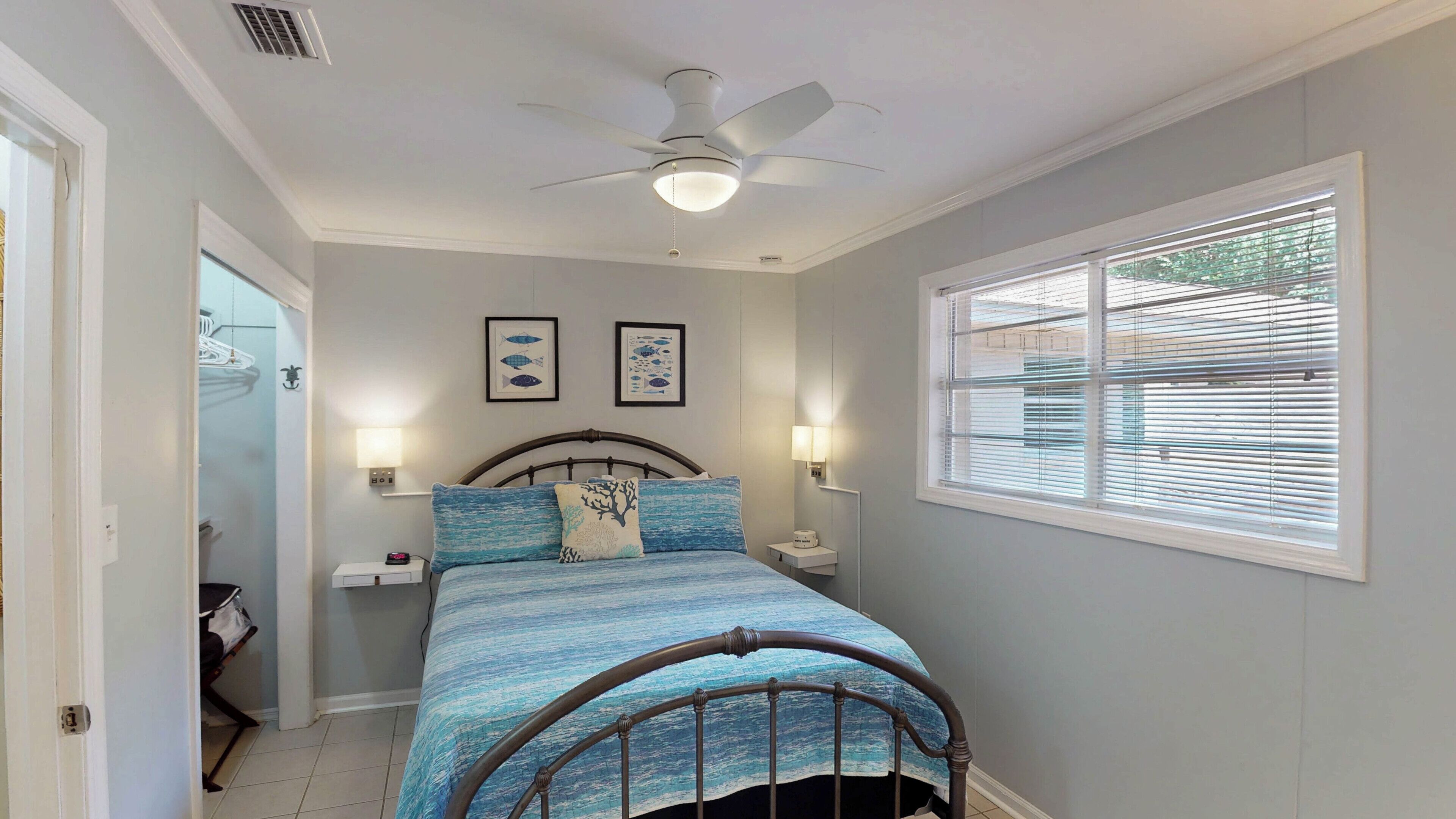 A bedroom with a bed, a ceiling fan, two wall-mounted lamps, a window with blinds, and a door leading to another room.