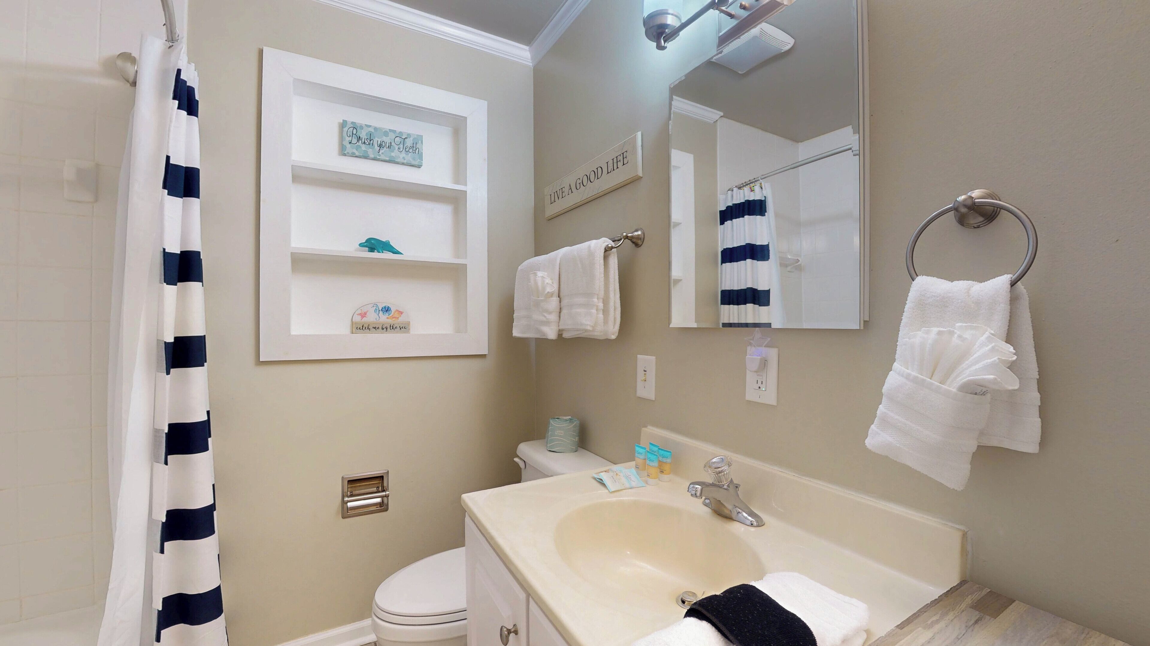 A bathroom with a toilet, sink, and shower curtain. There are towels hanging on the wall and a shelf with decorative items.